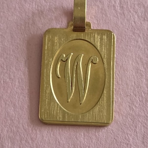 Op de afbeelding: Een rechthoekige goudkleurige hanger met een geborstelde metalen afwerking en een centrale ovale inkeping. De letter W is in cursief schrift gegraveerd in het ovaal. Een kleine, platte rechthoekige hangerlus is aan de bovenkant bevestigd.