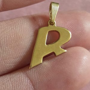 Może przedstawiać: Złoty metalowy wisiorek w kształcie wielkiej litery R. Wisiorek ma gładką, polerowaną powierzchnię i posiada teksturowany, prostokątny uchwyt u góry, służący do zawieszenia go na łańcuszku lub sznurku naszyjnika.