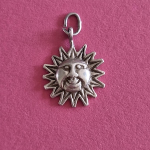 Op de afbeelding: Een zilverkleurige metalen hanger in de vorm van een zon met een centraal gezicht met ogen, een neus en een glimlachende mond. De zon heeft meerdere puntige stralen die vanuit het ronde midden naar buiten steken en een kleine ring bovenaan.