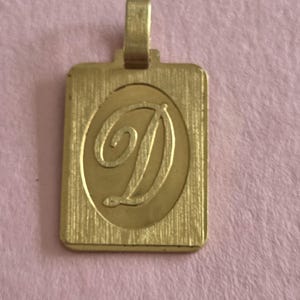 Op de afbeelding: Een rechthoekige goudkleurige hanger met een geborstelde metaaltextuur op het oppervlak. Het midden van de hanger bevat een gladde, verhoogde ovale vorm met een grote, cursieve letter D in het midden gegraveerd. Een klein oogje zit bovenaan.