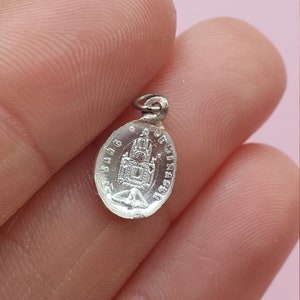 May include: Silver oval charm with a detailed image of a church and the text "S.S.A.G. DE FIRACRUCE".