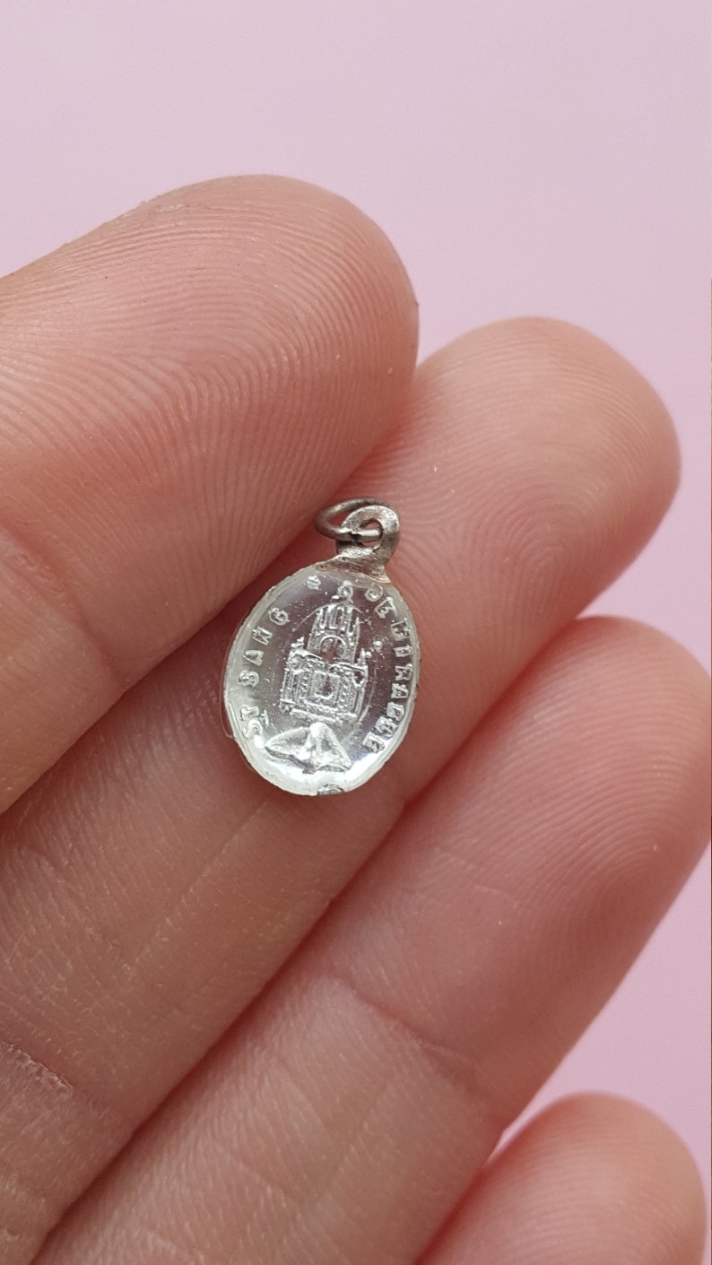 May include: Silver oval charm with a detailed image of a church and the text "S.S.A.G. DE FIRACRUCE".