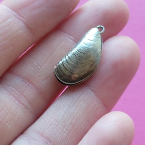 Könnte beinhalten: Ein kleiner, silberfarbener Anhänger in Form einer Muschelschale. Der Anhänger hat eine strukturierte Oberfläche mit detaillierten Rippen und eine kleine Schlaufe oben zum Befestigen an einer Kette oder einem Armband. Der Hintergrund ist einfarbig rosa.
