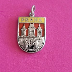 Könnte beinhalten: Ein schildförmiger Anhänger aus silberfarbenem Metall mit rotem Hintergrund und einer erhabenen silbernen Darstellung einer Burgmauer mit drei Türmen. Der obere Teil ist ein gelber horizontaler Balken mit dem Wort PRAHA in silbernen Buchstaben.