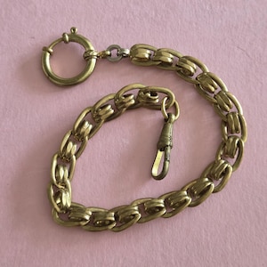 May include: A gold-coloured metal watch chain featuring a series of interlocking double links. The chain includes a circular spring ring clasp at one end and a small, elongated swivel hook clasp at the other, both designed for securing a pocket watch.