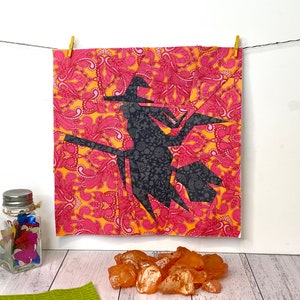 May include: A black paper cutout of a witch flying on a broomstick, against a pink and orange floral patterned fabric background. The fabric is hanging on a clothesline with wooden clothespins.