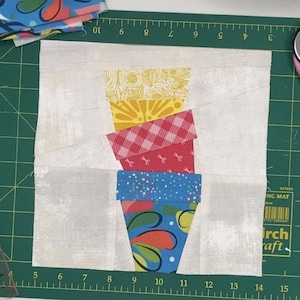 Puede incluir: Un cuadrado de tela blanco con una pila de cuatro cuadrados de tela en forma de cono de helado. Los cuadrados son amarillos con un patrón floral, rosa con un patrón de cuadros, rojo con un patrón de tijeras y azul con un patrón floral.