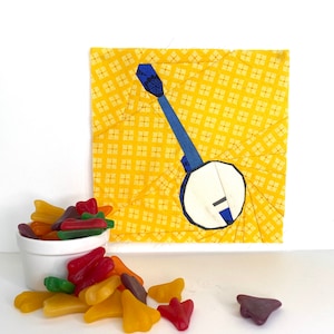 Puede incluir: Un cuadrado de tela amarillo con un diseño de banjo azul y blanco. Un tazón blanco con caramelos coloridos con forma de alas de avión está en primer plano.