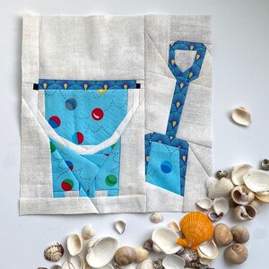 Peut inclure: Un seau et une pelle en tissu bleu et blanc avec un tissu à motifs bleus, blancs et rouges à l'intérieur. Le seau et la pelle sont sur un fond blanc avec un tas de coquillages.