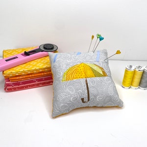 May include: A grey pincushion with a yellow and brown patchwork umbrella design. The pincushion has a pocket for storing pins and needles. There are several colourful pins stuck in the pincushion. Three spools of thread are on the table, one grey and two yellow.