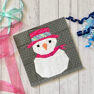 Peut inclure: Un bonhomme de neige blanc avec un chapeau et une écharpe roses, des yeux noirs et un nez orange, sur un fond gris à pois blancs.