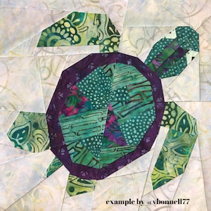 May include: A green and purple patchwork quilt block depicting a turtle. The turtle's shell is made of various green and purple fabrics with a purple border. The turtle's head and legs are also made of patchwork fabric.