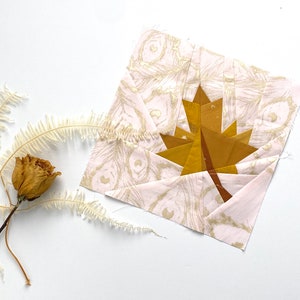 Pode incluir: Um bloco de tecido com um desenho de folha de bordo dourada. A folha é feita de tecido dourado. O tecido de fundo é rosa claro com um padrão de penas douradas. Uma rosa e uma samambaia secas também estão na imagem.