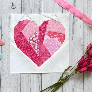 May include: A pink and white patchwork heart quilt block with a white background. The heart is made of various pink and red fabrics with different patterns.