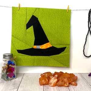 May include: A green fabric witch hat with an orange and black trim hangs on a clothesline. The hat is made of fabric squares sewn together. A black beaded necklace with purple beads and a clear crystal pendant hangs on the right side of the image.