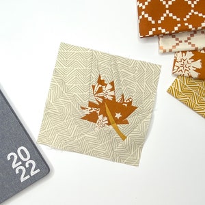 May include: A square fabric patch with a brown maple leaf design, featuring white stars and snowflake accents, sits on a white surface. A grey 2022 notebook and several patterned fabric swatches in brown and white are also visible.