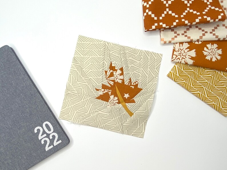 May include: A square fabric patch with a brown maple leaf design, featuring white stars and snowflake accents, sits on a white surface. A grey 2022 notebook and several patterned fabric swatches in brown and white are also visible.