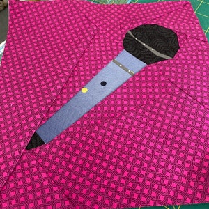 May include: A fabric square featuring a microphone applique. The microphone has a black head and a blue body with yellow and black accents. The background is a magenta fabric with a repeating black circular pattern, creating a bold, graphic design.