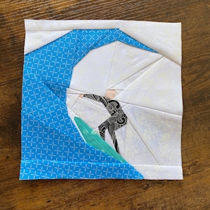 May include: A blue and white fabric patchwork quilt block depicting a surfer riding a wave. The surfer is wearing a black wetsuit with white accents and a black and white pattern. The wave is a light blue and white fabric with a white background.