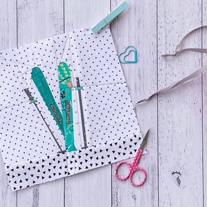 Peut inclure: Un bloc de tissu blanc avec un motif de pois noirs et un motif de triangles noirs et blancs sur le bord inférieur. Le bloc est plié en deux et présente un motif de tissu vert et blanc à l'intérieur. Il y a des ciseaux roses, un trombone en forme de cœur bleu et un sac en tissu rose avec un cordon sur une surface en bois blanc.