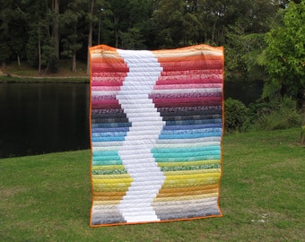 May include: A colorful quilt with a white X pattern in the center. The quilt is made of various fabrics in shades of red, orange, yellow, green, blue, and purple. The quilt is displayed outdoors on a grassy lawn.