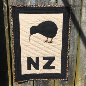 Peut inclure: Une tenture murale en tissu beige avec une silhouette d'oiseau kiwi noir et les lettres NZ en noir. Le tissu a une bordure à motifs noirs et dorés.