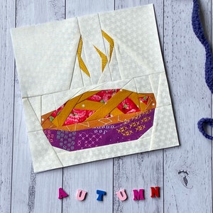 May include: A patchwork quilt block featuring a pie with a purple crust, a golden lattice top, and red filling. The block is surrounded by a white background with a small, gray pattern. The word "AUTUMN" is spelled out in colorful letters below the block.