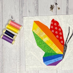 Peut inclure: Un bloc de courtepointe en forme de papillon aux couleurs de l'arc-en-ciel sur fond blanc. Le papillon a une aile rouge à pois blancs et une aile verte à rayures jaunes. Le papillon est fait de chutes de tissu et est un excellent exemple de bloc de courtepointe en patchwork.