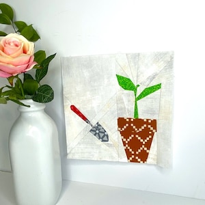 May include: A white background with a green plant in a brown pot with a red and gray gardening tool. The plant has two green leaves and is in a brown pot with a geometric pattern.