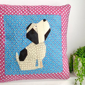 May include: A pink and white polka dot pillow with a blue and white patterned square in the center. The square features a black and white geometric dog design.