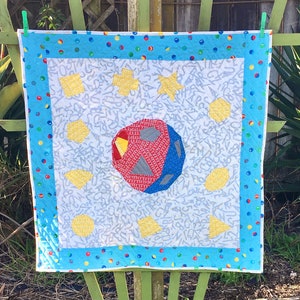 May include: A white baby quilt with a blue, red, and yellow geometric design. The quilt has a blue border with white polka dots.