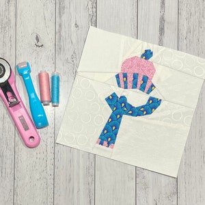 May include: A white fabric square with a blue and pink applique of a snowman wearing a hat and scarf. The fabric square is surrounded by a white wooden surface with three spools of thread, a rotary cutter, and a ruler.