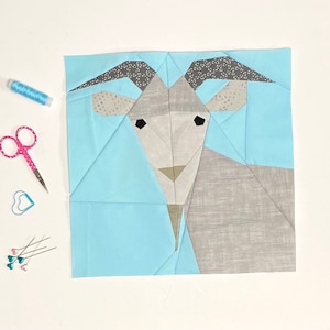 Op de afbeelding: Een lichtblauw vierkant van stof met een geometrisch geitenontwerp in grijs en wit. De geit heeft zwarte ogen en een bruine neus.