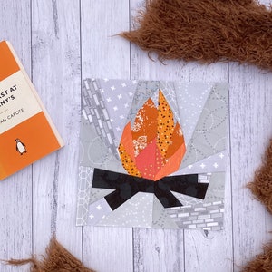 May include: A patchwork quilt block featuring a stylised campfire with orange, red and black fabric pieces. The background is a light grey fabric with white and grey geometric patterns.
