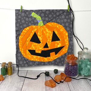 May include: A patchwork pumpkin with a jack-o'-lantern face, made from orange, yellow, and black fabric. The pumpkin is hanging from a string and is surrounded by small jars of glitter and colorful stones.