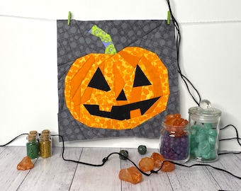 Op de afbeelding: Een patchwork-pompoen met een jack-o'-lantern-gezicht, gemaakt van oranje, gele en zwarte stof. De pompoen hangt aan een touwtje en is omringd door kleine potjes met glitter en gekleurde stenen.