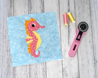 Può includere: Un cavalluccio marino in carta tagliata rosa e giallo su uno sfondo blu. Il cavalluccio marino è fatto di ritagli di tessuto ed è circondato da cuciture bianche. Lo sfondo è un tessuto blu chiaro con un motivo a marmo bianco.