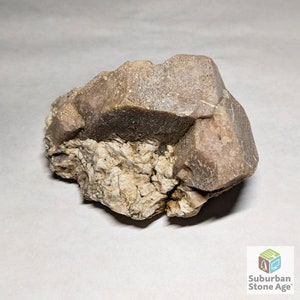 Puede incluir: Primer plano de una roca marrón y blanca con un gran cristal marrón rugoso. El cristal tiene un tono ligeramente rosado y está rodeado de cristales blancos más pequeños.