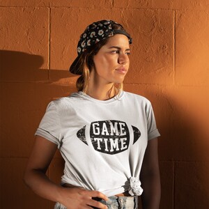Puede incluir: Una mujer con una camiseta blanca con el texto "GAME TIME" impreso en negro con una fuente desgastada dentro de una forma de bal&oacute;n de f&uacute;tbol. Lleva una gorra de b&eacute;isbol con estampado floral y pantalones cortos de mezclilla.