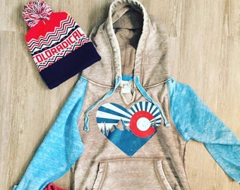 Könnte beinhalten: Ein brauner und blauer Hoodie mit einem herzförmigen Grafik, das eine Bergkette und einen roten Kreis mit weißem Umriss zeigt. Der Hoodie hat eine blaue und weiße Bommelmütze und rote Fäustlinge.