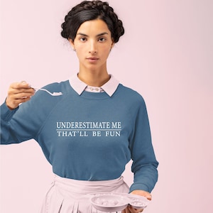May include: A woman wearing a blue sweatshirt with the text "UNDERESTIMATE ME THAT'LL BE FUN" in white. She is holding a white spoon in her right hand and a white plate in her left hand.