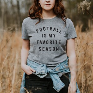 Puede incluir: Una mujer con una camiseta gris con el texto "Football is my favorite season" impreso en ella. Est&aacute; de pie en un campo de hierba alta.