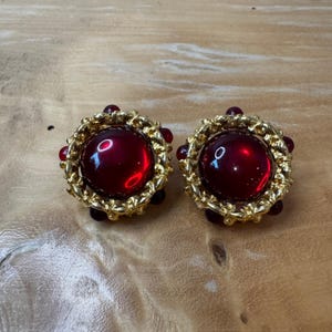 May include: A pair of vintage clip-on earrings. Each earring features a large, round, deep red glass stone surrounded by a gold-toned metal filigree. Small red beads accent the outer edge of the filigree, creating a striking contrast.