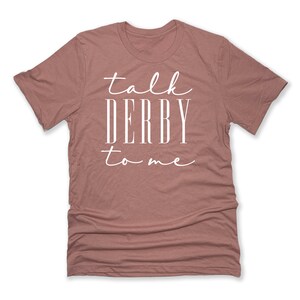 Puede incluir: Camiseta rosa polvoriento con la frase blanca "talk DERBY to me". El texto es una mezcla de escritura y letras de bloque. La camiseta tiene cuello redondo y est&aacute; hecha de un material suave. El texto est&aacute; centrado.