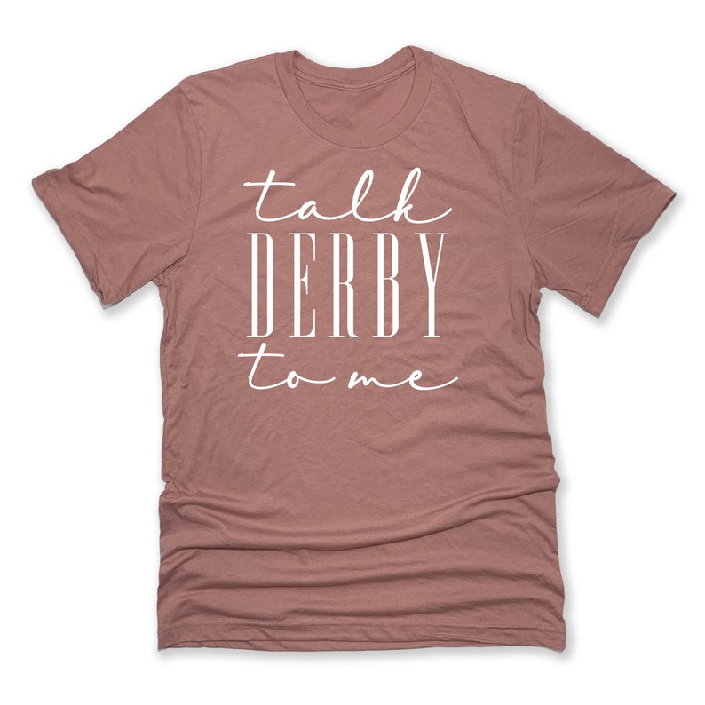 Puede incluir: Camiseta rosa polvoriento con la frase blanca "talk DERBY to me". El texto es una mezcla de escritura y letras de bloque. La camiseta tiene cuello redondo y est&aacute; hecha de un material suave. El texto est&aacute; centrado.