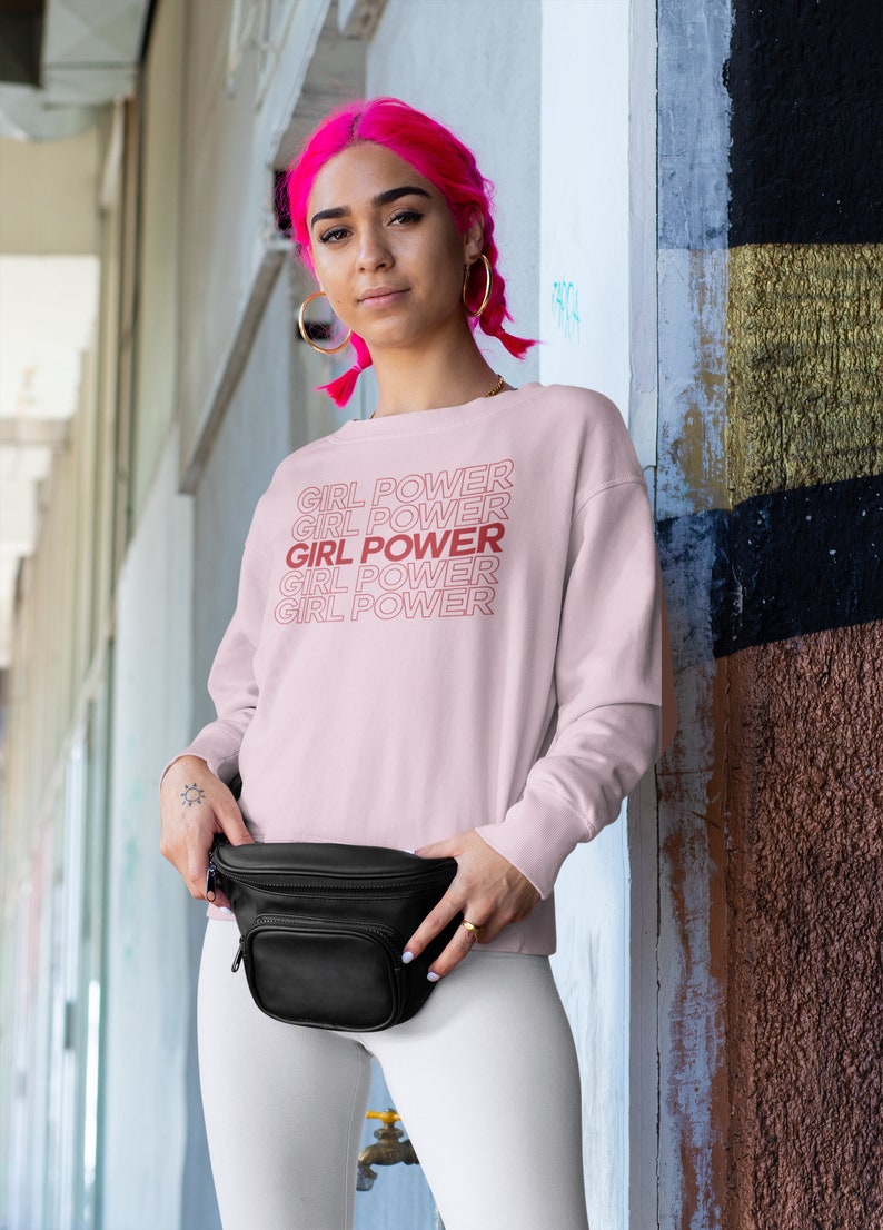May include: A pink sweatshirt with the text "GIRL POWER" repeated four times in red. The model is wearing white pants and a black fanny pack.