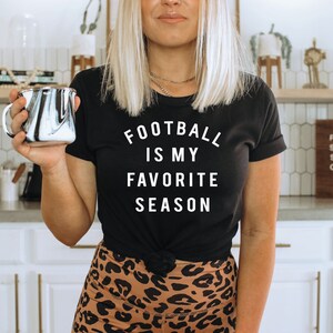 Puede incluir: Una mujer lleva una camiseta negra con el texto "Football is my favorite season" en blanco. Est&aacute; sosteniendo una taza marmoleada en blanco y negro. Est&aacute; de pie en una cocina con gabinetes blancos y un letrero de madera que dice "Pour some sugar on me - Coffee".