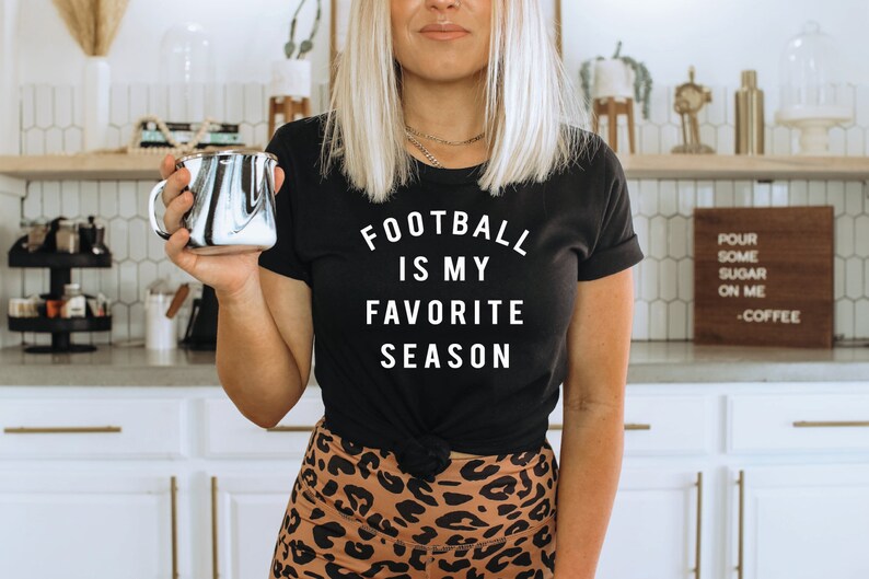 Puede incluir: Una mujer lleva una camiseta negra con el texto "Football is my favorite season" en blanco. Est&aacute; sosteniendo una taza marmoleada en blanco y negro. Est&aacute; de pie en una cocina con gabinetes blancos y un letrero de madera que dice "Pour some sugar on me - Coffee".