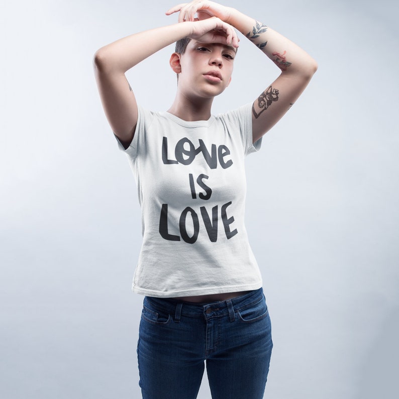 May include: A person wearing a white t-shirt with the text "LOVE IS LOVE" in black lettering. The person is wearing blue jeans and has tattoos on their arms.