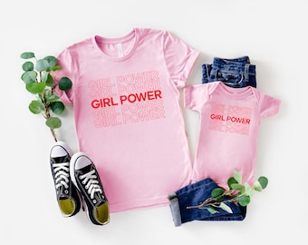 Könnte beinhalten: Rosa T-Shirts und ein Baby-Body mit dem Text "GIRL POWER" in Rot und Rosa. Das Bild enthält ein Paar schwarze Turnschuhe, gefaltete blaue Jeans und Grünzeug.